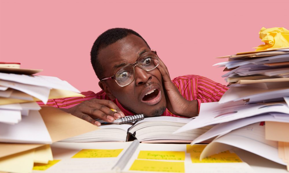 Desperate nervous dark skinned male student looks stressfully at pile of papers, writes information in notepad, has much work and deadline for preparation, isolated on pink wall. Close up shot