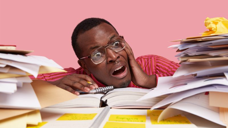 Desperate nervous dark skinned male student looks stressfully at pile of papers, writes information in notepad, has much work and deadline for preparation, isolated on pink wall. Close up shot